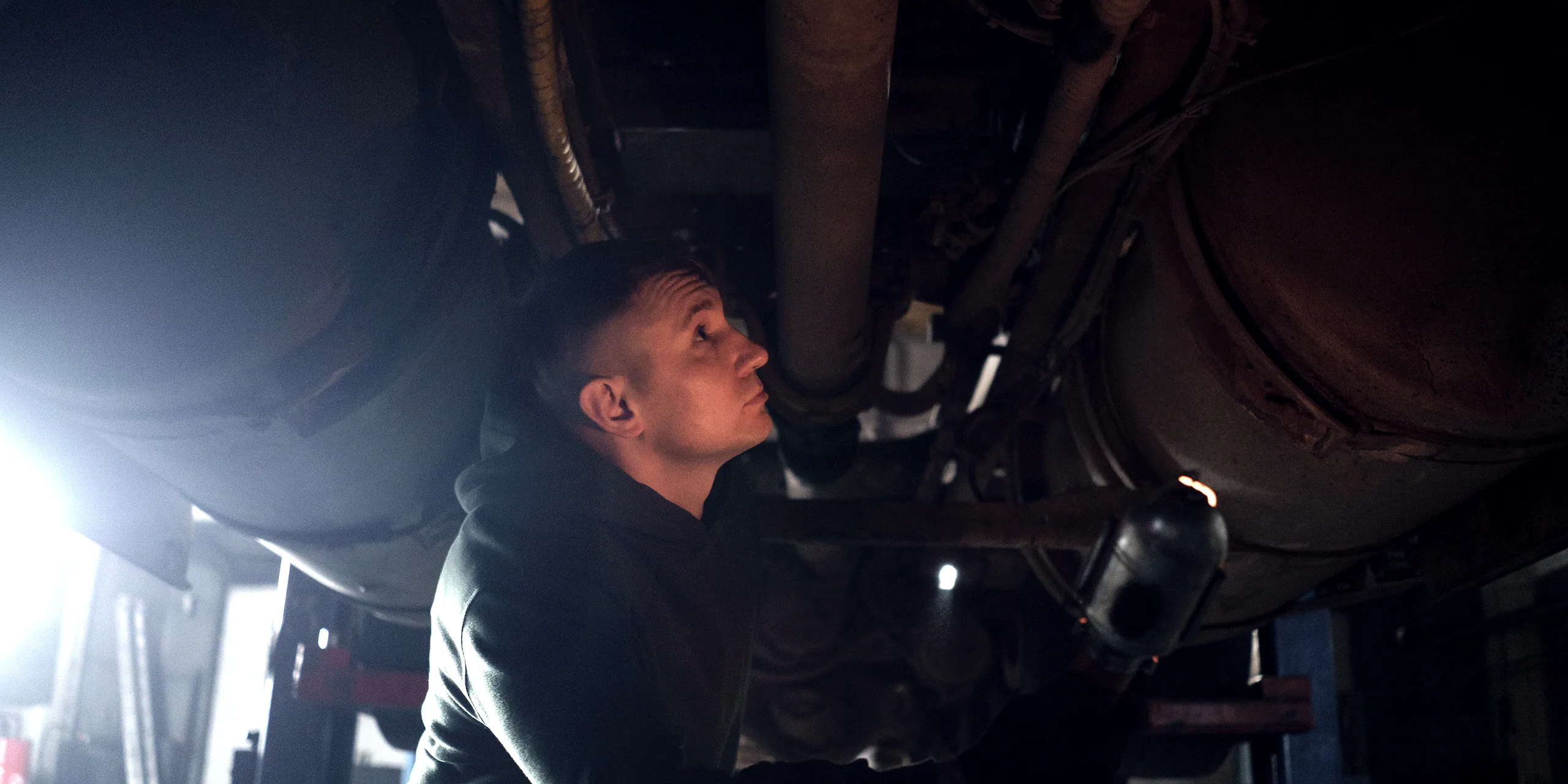 A technician working underneath a car on a lift, lit by a hand lamp.