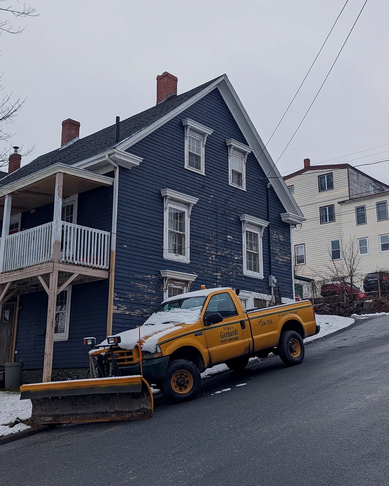 A pickup truck with a plow blade parked on a snowy street in front of a clapboard house.