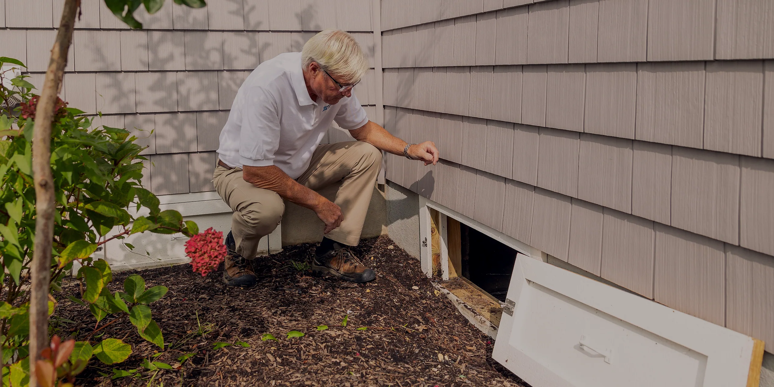 A technician crouched at an open crawl space access on the side of a clapboard house.