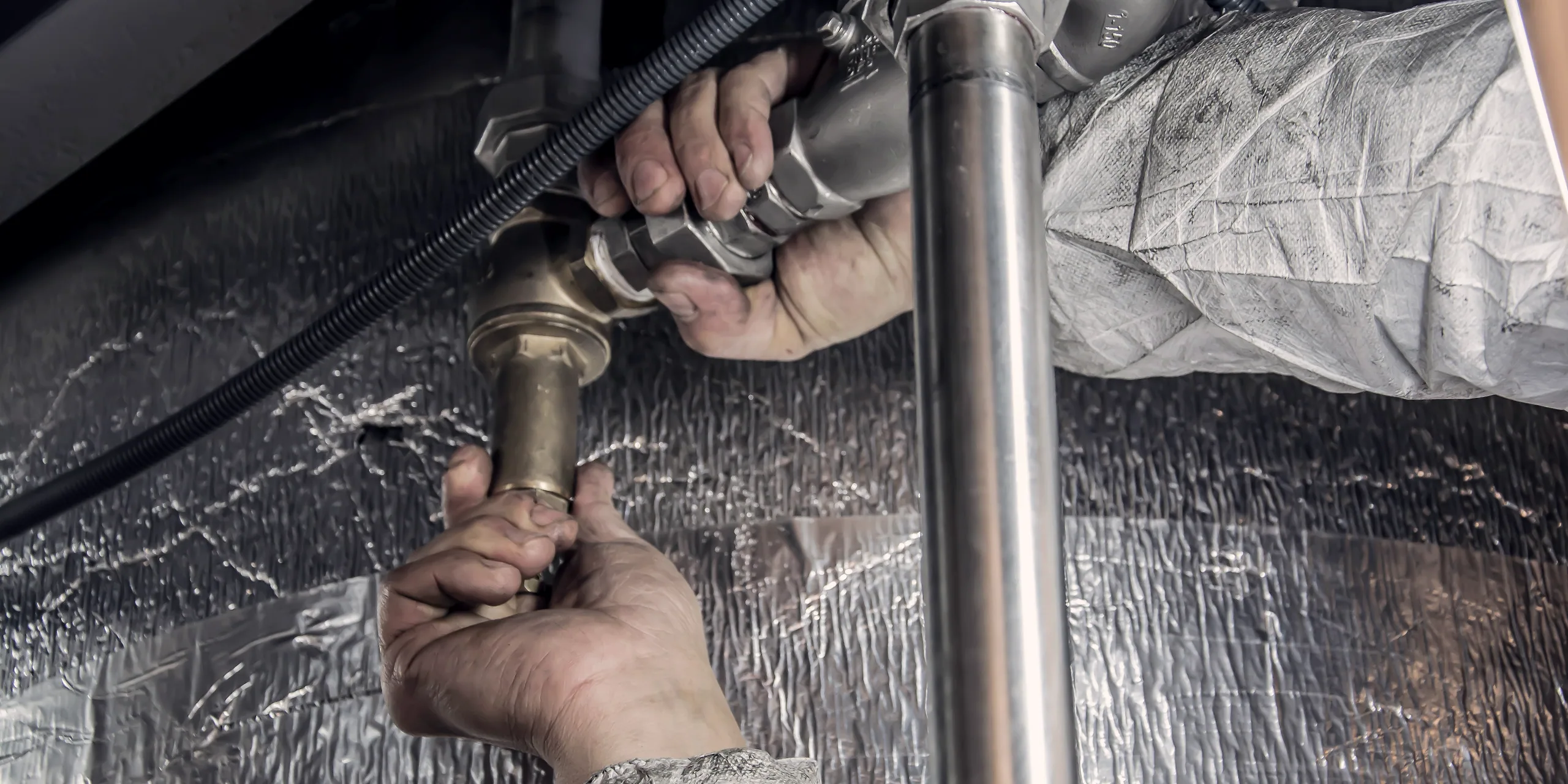 A plumber's hands tightening a fitting on a copper line under insulated pipework.