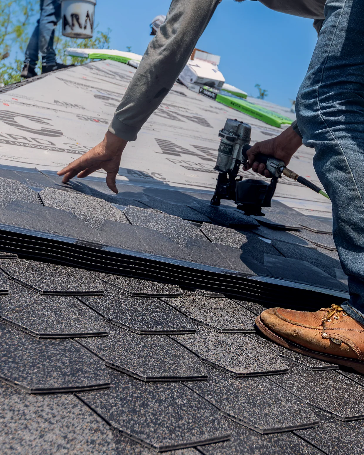 A roofer's hands setting an asphalt shingle, nail gun in the other hand.